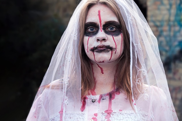 Retrato de mujer zombie en vestido de novia con velo y maquillaje de escenario mirando a la chica de la cámara aplicar