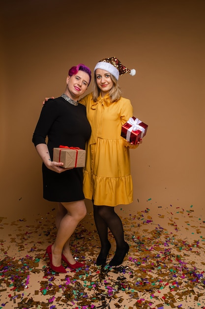 Retrato de mujeres alegres intercambiando regalos de Navidad y sonriendo. Rubia con sombrero de Santa brillante y vestido amarillo