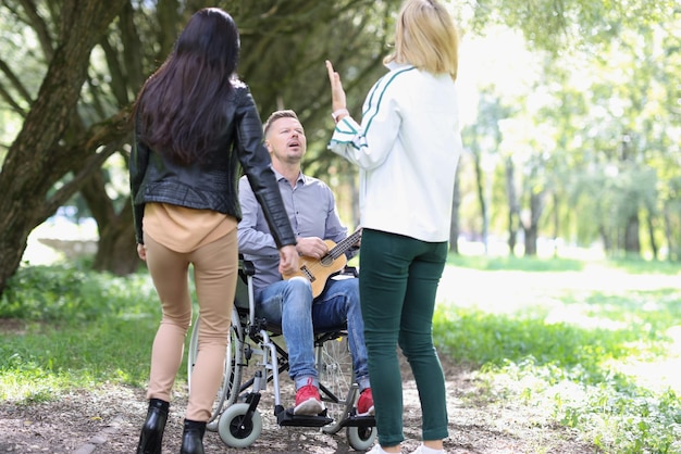 Retrato de mujeres divirtiéndose con un hombre discapacitado en silla de ruedas en un chico del parque jugando con el ukelele