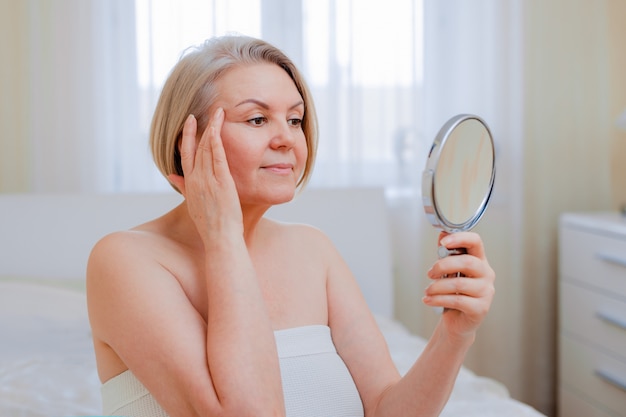 Retrato mulher muito sênior com as mãos no espelho do rosto em casa depois do banheiro Cuidados com a pele após 50-60 anos