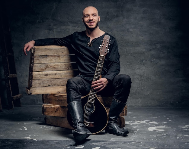 Retrato de un músico masculino de cabeza rapada vestido con ropa celta vieja se sienta en una caja de madera y toca mandolina vieja.