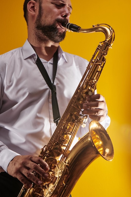 Retrato de músico profesional saxofonista con camisa blanca toca música de jazz en saxofón, fondo amarillo en un estudio fotográfico, vista lateral