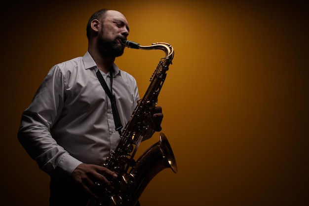 Retrato de músico profesional saxofonista con camisa blanca toca música de jazz en saxofón, fondo amarillo en un estudio fotográfico, vista lateral