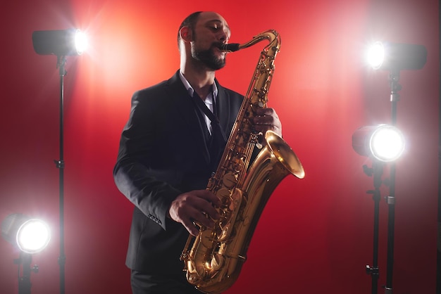 Retrato de músico profesional saxofonista hombre en traje toca música jazz en saxofón, fondo rojo en un estudio fotográfico