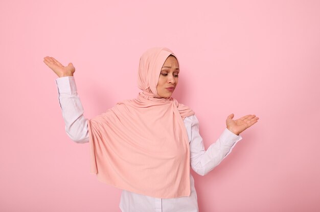 Retrato na altura da cintura de uma linda mulher muçulmana misteriosa usando um lenço na cabeça hijab, mostrando algo nas palmas das mãos, isolado em um fundo rosa colorido, com espaço de cópia para publicidade ou promoção