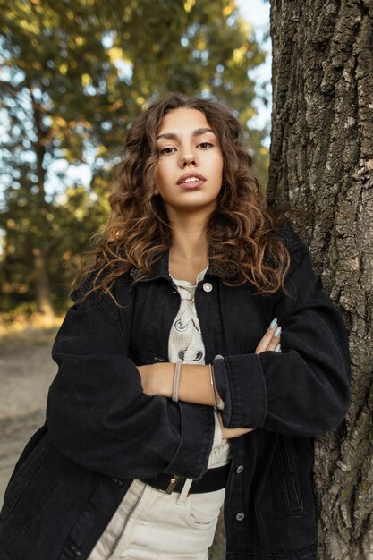 Retrato natural de una bella mujer joven con cabello rizado en ropa de mezclilla de moda se encuentra cerca de un árbol en el campo. Belleza y estilo de jeans femeninos