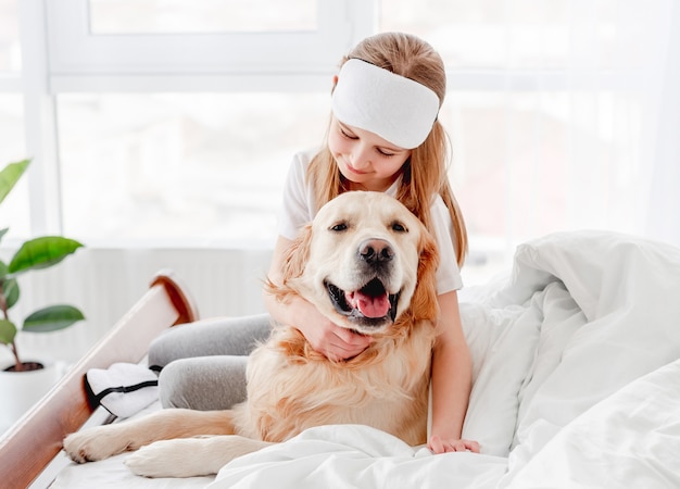 Retrato de niña abrazando a perro golden retriever en la cama después de despertarse. Niño que se queda en casa con un perrito por la mañana y lo acaricia. Animal doméstico con dueño