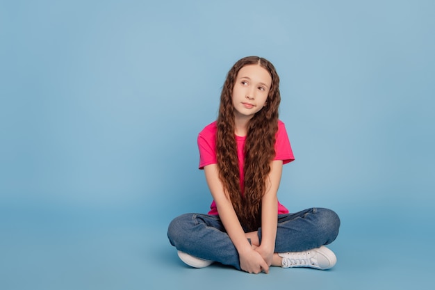 Retrato de niña adorable interesada sentarse en el suelo buscar un espacio en blanco sobre fondo azul.