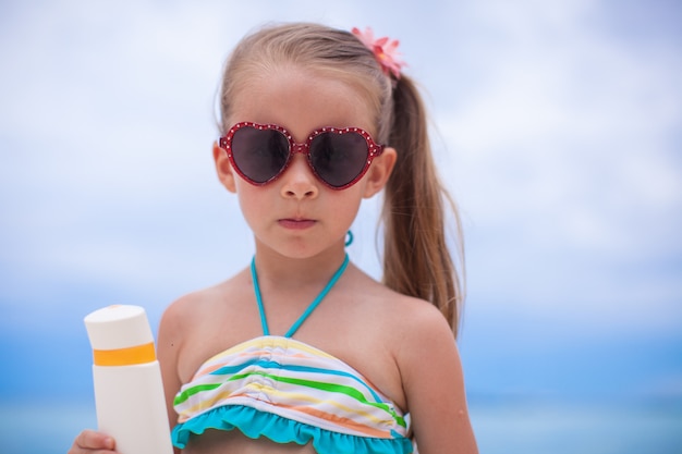 Retrato de niña adorable en traje de baño tiene botella de loción bronceadora