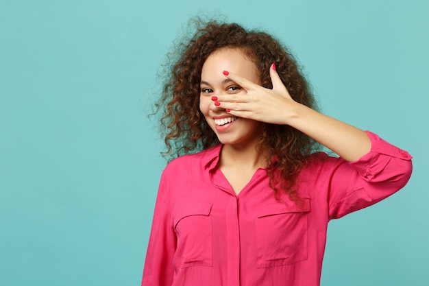 Retrato de niña africana alegre en ropa casual rosa que cubre la cara con la mano aislada sobre fondo de pared azul turquesa en estudio. Personas sinceras emociones, concepto de estilo de vida. Simulacros de espacio de copia.