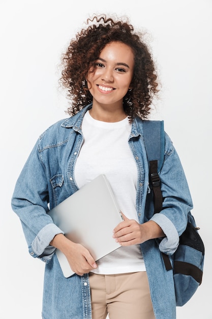 Retrato de una niña africana casual bastante alegre que lleva la mochila que se encuentran aisladas sobre la pared blanca, sosteniendo la computadora portátil