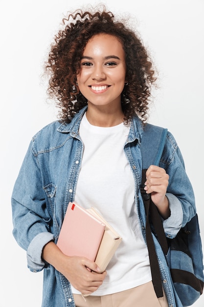 Retrato de una niña africana casual bastante alegre que lleva la mochila que se encuentran aisladas sobre la pared blanca, sosteniendo libros de texto