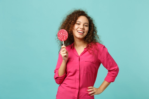 Retrato de niña africana encantadora en ropa casual sosteniendo en la mano piruleta redonda rosa aislada sobre fondo azul turquesa en estudio. Personas sinceras emociones, concepto de estilo de vida. Simulacros de espacio de copia.