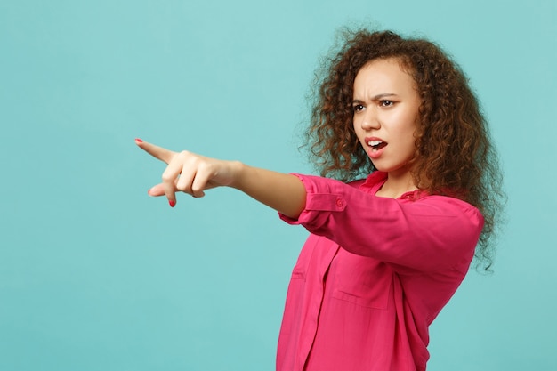 Retrato de niña africana irritada en ropa casual jurando, apuntando con el dedo índice a un lado aislado sobre fondo de pared azul turquesa. Personas sinceras emociones, concepto de estilo de vida. Simulacros de espacio de copia.