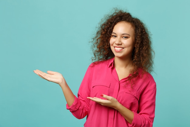 Retrato de niña africana linda sonriente en ropa casual rosa apuntando con las manos a un lado aislado sobre fondo de pared azul turquesa en estudio. Personas sinceras emociones, concepto de estilo de vida. Simulacros de espacio de copia.