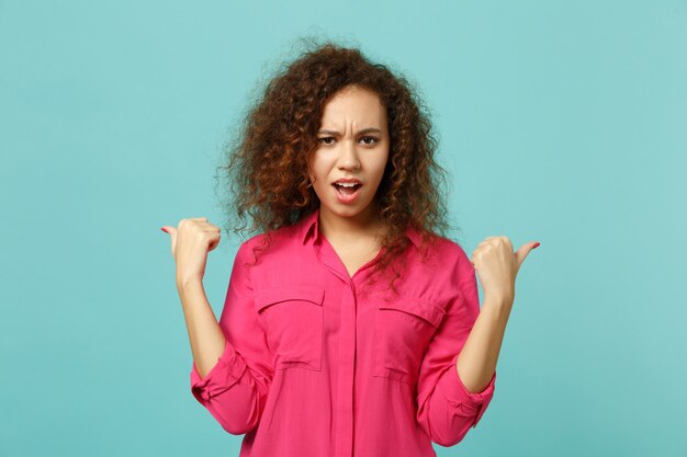 Retrato de niña africana perpleja en ropa casual rosa apuntando los pulgares a un lado aislado sobre fondo de pared azul turquesa en estudio. Concepto de estilo de vida de emociones sinceras de personas. Simulacros de espacio de copia.