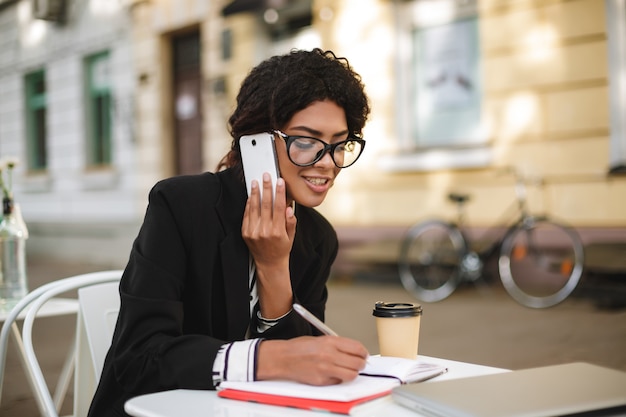 Retrato de niña afroamericana sentada en la mesa del café y escribiendo en su cuaderno mientras habla por teléfono celular