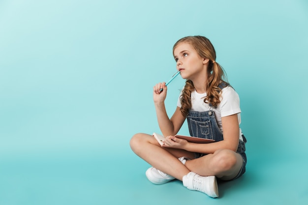 Retrato de una niña alegre aislada sobre pared azul, escribiendo en un cuaderno