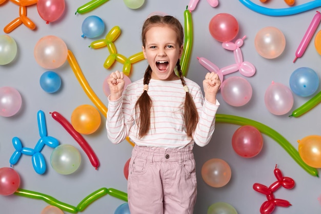 Retrato de una niña alegre y alegre con trenzas con ropa informal gritando de felicidad celebrando el cumpleaños de pie contra una pared gris con globos de colores