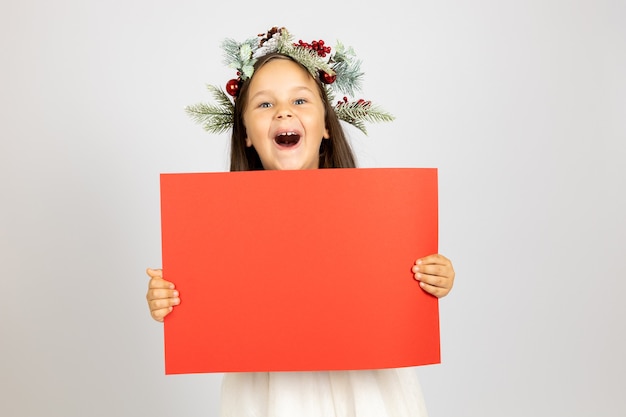 Retrato de niña alegre riendo en corona de Navidad sosteniendo pancarta roja aislado sobre fondo blanco.