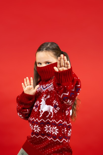 Retrato de niña alegre en suéter rojo de navidad con renos y con cuello en media cara mostrando ...