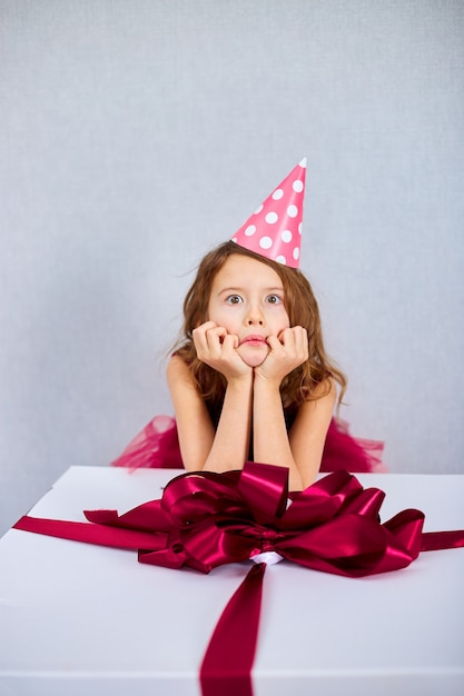 Retrato de niña alegre en vestido rosa y sombrero puso sus codos en caja de regalo grande