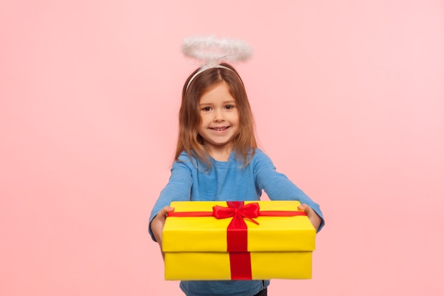 Foto retrato de niña angelical amable generosa con nimbus dando gran regalo de cumpleaños a la cámara y sonriendo, felicitando por vacaciones, compartiendo regalo. foto de estudio de interior aislado sobre fondo rosa