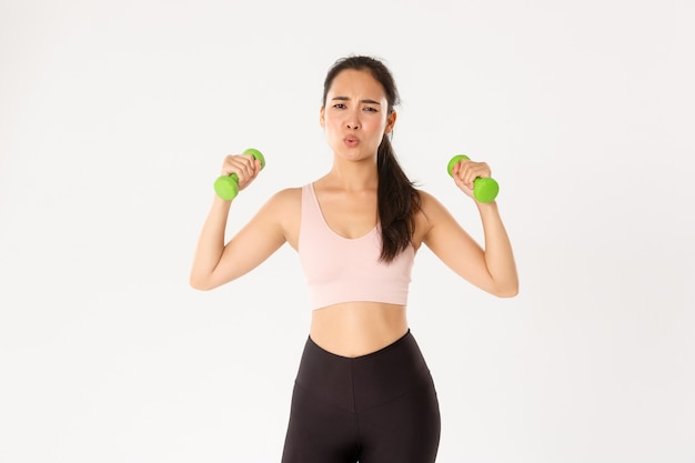 Retrato de niña asiática cansada en ropa deportiva, luciendo agotada durante el entrenamiento, ejercicio en casa con el entrenador en línea, levantando pesas, fondo blanco.