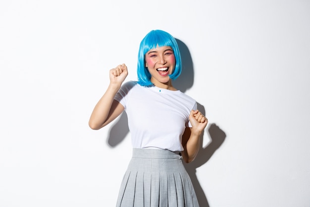 Retrato de niña asiática despreocupada con peluca azul divirtiéndose en la fiesta, celebrando halloween, bailando y riendo, de pie.