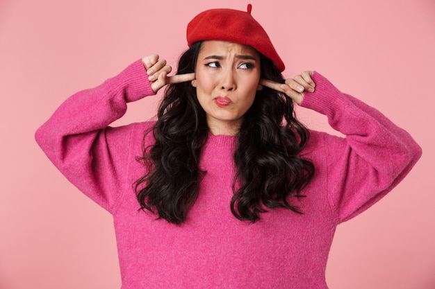 Retrato de niña asiática hermosa molesta con cabello largo oscuro con boina tapándose los oídos con los dedos aislados en rosa