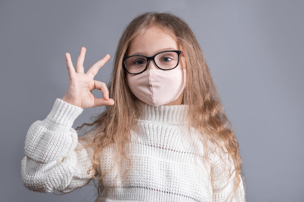 Retrato de una niña atractiva joven con cabello largo y rubio que fluye en un suéter blanco en máscara protectora médica muestra signo bien