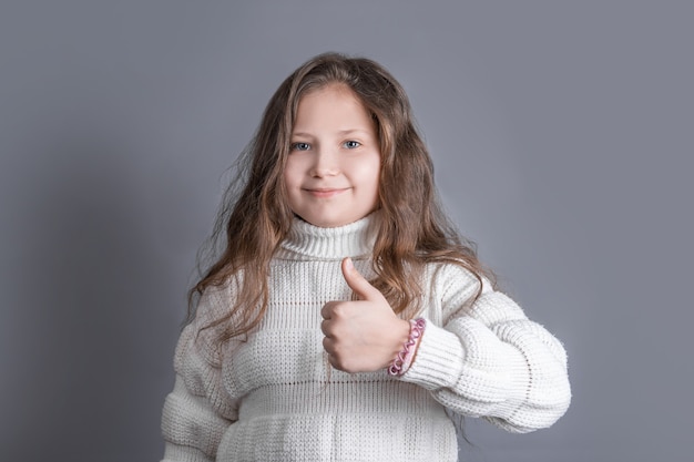 Retrato de una niña atractiva joven con cabello largo y rubio que fluye con un suéter blanco sonriendo muestra los pulgares hacia arriba, como cantar sobre un fondo gris de estudio. Lugar para el texto. Copie el espacio.