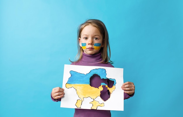 Retrato de una niña con una bandera ucraniana pintada en la cara de pie sobre un fondo azul y sosteniendo un mapa quemado de Ucrania en sus manos Niños de Ucrania contra la guerra
