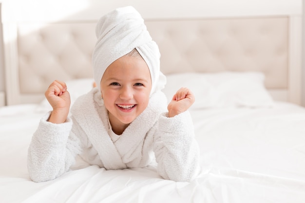 Retrato de una niña en una bata de baño blanca con una toalla blanca en la cabeza acostada en la cama