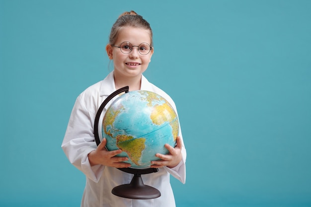 Retrato de niña en bata blanca sosteniendo el mapa del mundo y sonriendo a la cámara contra el fondo azul.