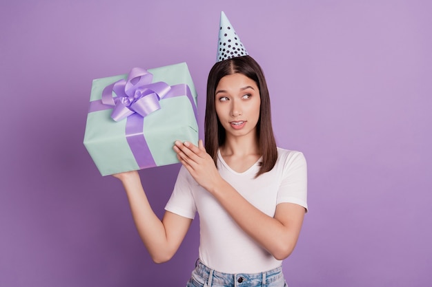 Retrato de niña bonita con gorro de papel sosteniendo en las manos giftbox agitar mirada curiosa espacio vacío aislado sobre fondo de color violeta vivo