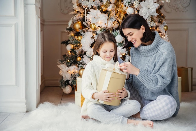 Retrato de una niña bonita y una madre hermosa sentadas juntas en una alfombra cálida, sostenga una caja de regalo, disfrute del árbol de Año Nuevo decorado. La familia pasa tiempo juntos Concepto de celebración y vacaciones