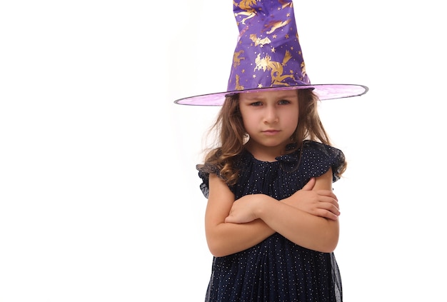 Retrato de niña bonita molesta enojada de bruja con un sombrero de mago y vestida con un elegante vestido de carnaval, mirando a la cámara posando con los brazos cruzados sobre fondo blanco, espacio de copia. Víspera de Todos los Santos