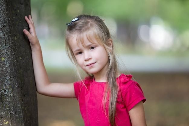 El retrato de una niña bonita del niño que se coloca cerca de tronco de árbol grande en verano parquea al aire libre.