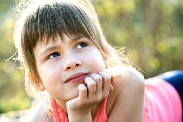 Retrato de niña bonita con ojos grises y cabello largo rubio apoyado en sus manos sonriendo felizmente al aire libre sobre fondo brillante borroso. Linda niña en un cálido día de verano afuera.