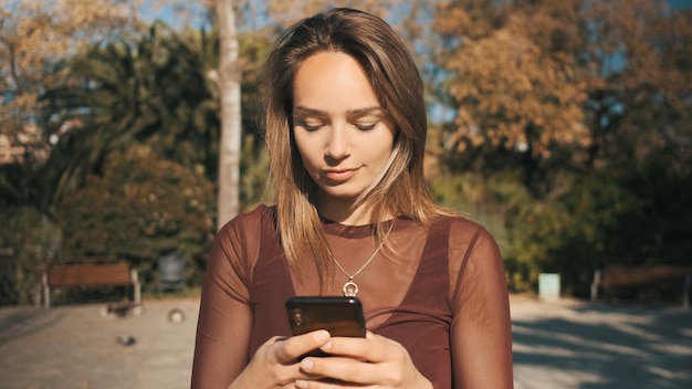 Retrato de niña bonita rubia con mensajes de texto de smartphone sensualmente con novio en el parque de la ciudad