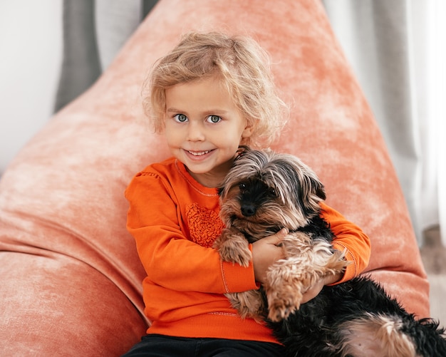 Retrato de niña bonita sentada en una silla naranja con un perro en sus brazos mirando a la cámara y sonriendo