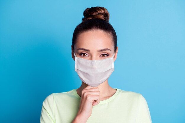 Retrato de niña bonita toque la barbilla del dedo mirar en la cámara use una máscara médica aislada sobre fondo de color azul