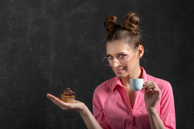 Retrato de niña bonita en vasos sosteniendo una taza de café y muffin de chocolate Mujer con peinado de moño Fondo negro