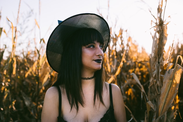 Retrato de niña bruja disfraz de Halloween en un maizal al atardecer. Hermosa mujer joven seria en sombrero de brujas con cabello largo y negro y labios oscuros