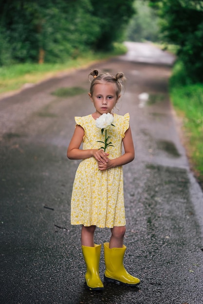 Retrato de niña caucásica de 6 años en vestido amarillo y botas de lluvia de pie en el parque con flor de peonía