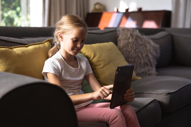 Retrato de una niña caucásica disfrutando de su tiempo en una casa, sentada en un sofá, usando una tableta digital, mirando la cámara, sonriendo y autoaislada en cuarentena durante el coronavirus covid