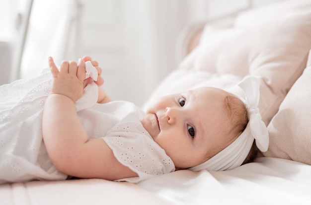 Retrato de una niña caucásica con un traje blanco y una banda para la cabeza acostada en una cama blanca en una habitación