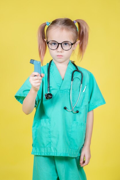 Foto retrato de niña caucásica vestida con abrigo verde de médicos con un inhalador para el asma en la mano sobre fondo amarillo