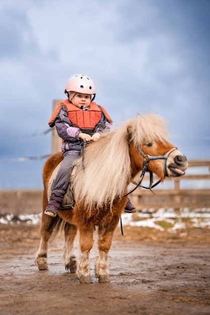 Retrato de niña con chaqueta protectora y casco con su pony marrón antes de la lección de equitación